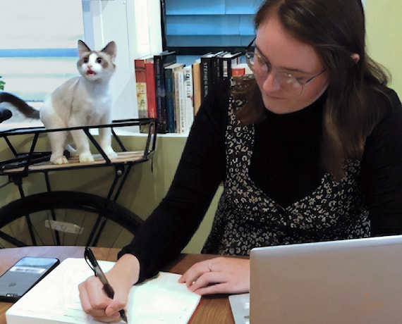 A young woman works at her desk in her home office. Her laptop is open in front of her, and she is writing on a notepad. Her cat in the background is mid-meow.
