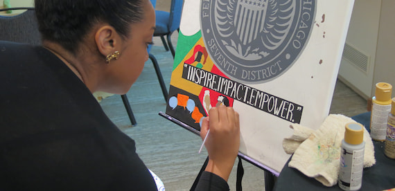 A woman paints a colorful canvas with the Black Impact Group's motto–Inspire, Impact, Empower–beneath the Seventh District seal.