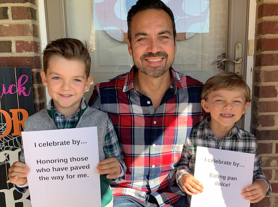 A father poses with his children. The children are holding signs that say, I celebrate by honoring those who have paved the way for me, and I celebrate by eating pan dulce!