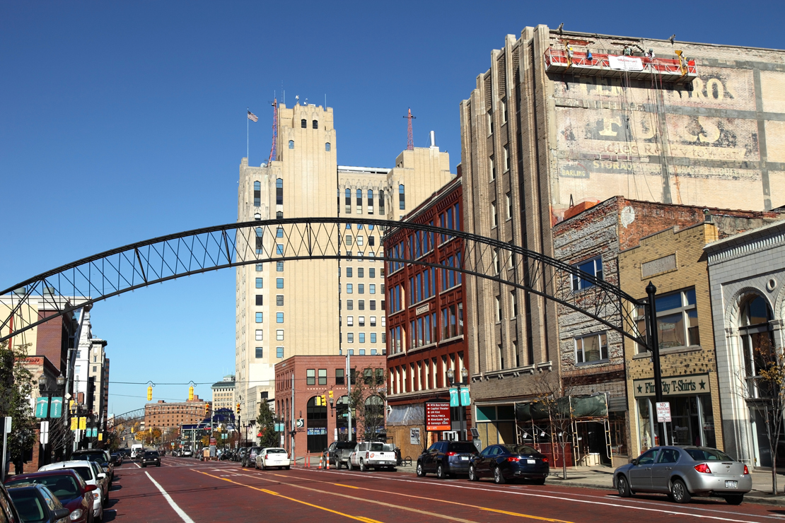 A color image of downtown Flint, Michigan during the daytime, featuring an iron arch that crosses the wide boulevard. The road looks to be made of red bricks.