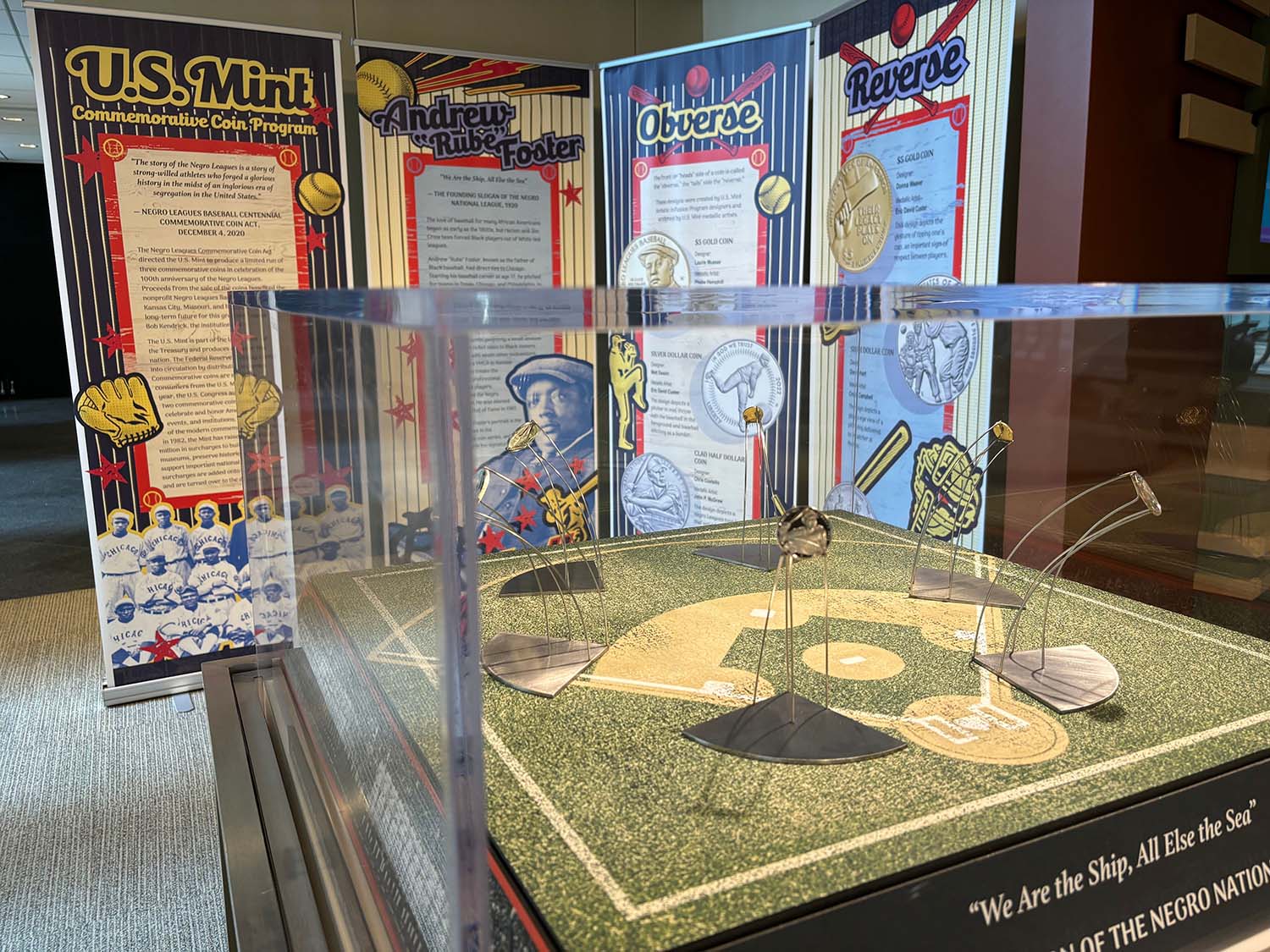 A display case holding commemorative coins featuring historic Black baseball players, on a base designed to look like a baseball diamond. In the background is a four-panel interpretive sign about the Negro Leagues Commemorative Coin Act and the images on the coins.