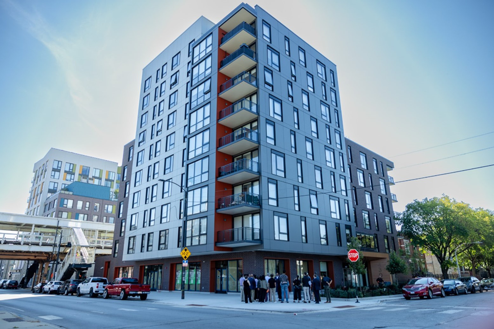 A group stands in front of an apartment building near elevated CTA tracks.