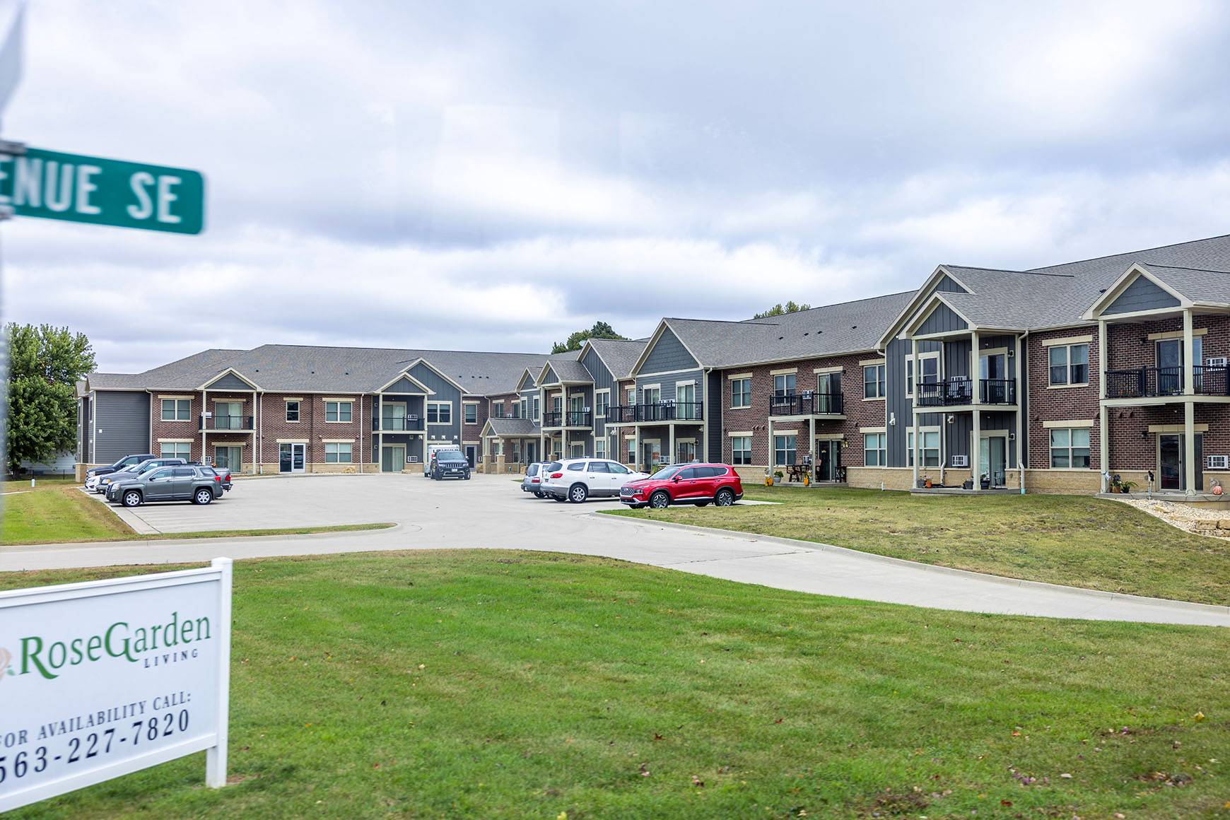 The picture features a two-story apartment building with a parking lot and a white sign saying “Rose Garden Living” in front.