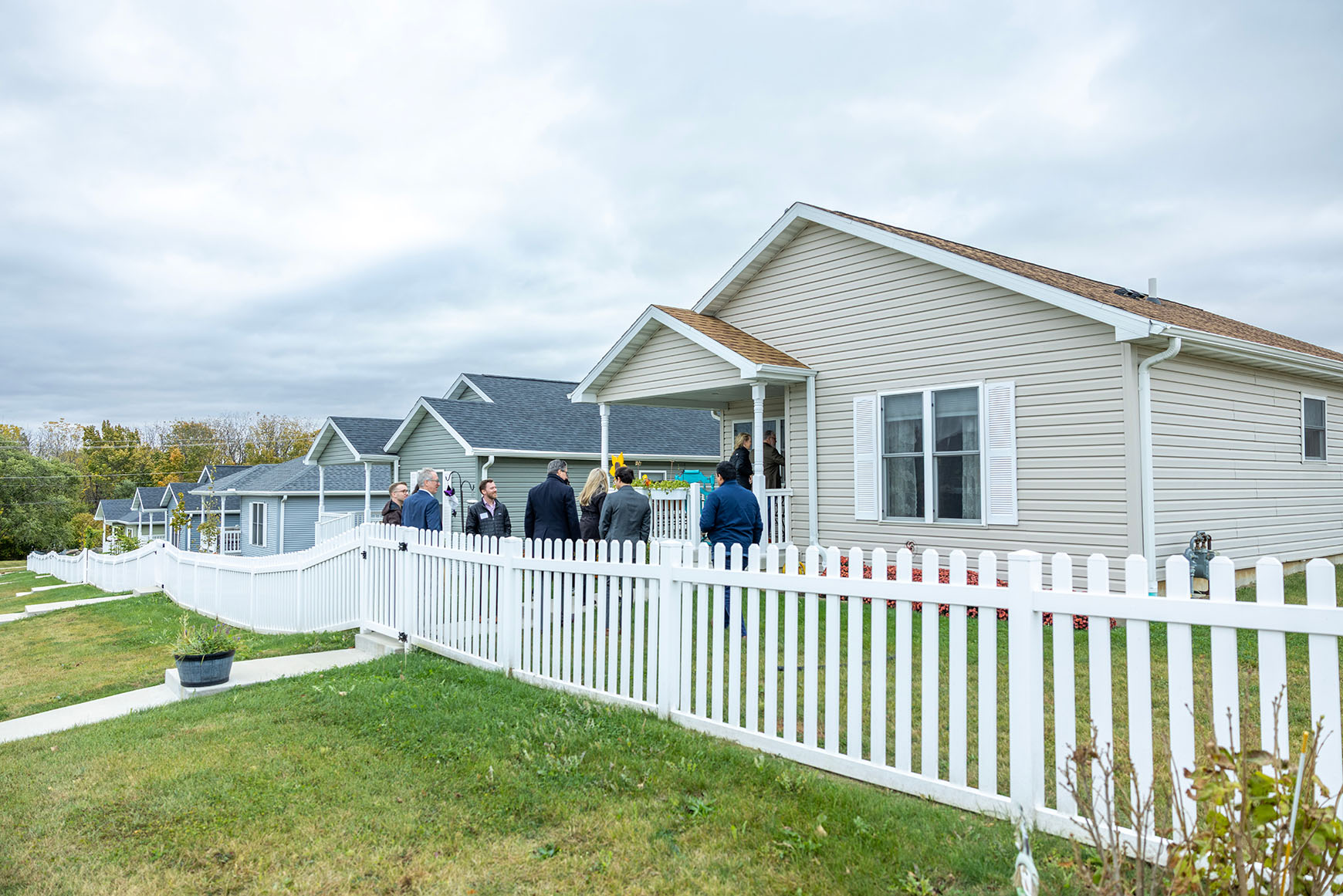Several tour participants stand in the front yard of a small single-family home. The home is situated in a row of similar houses, all enclosed by a white picket fence.