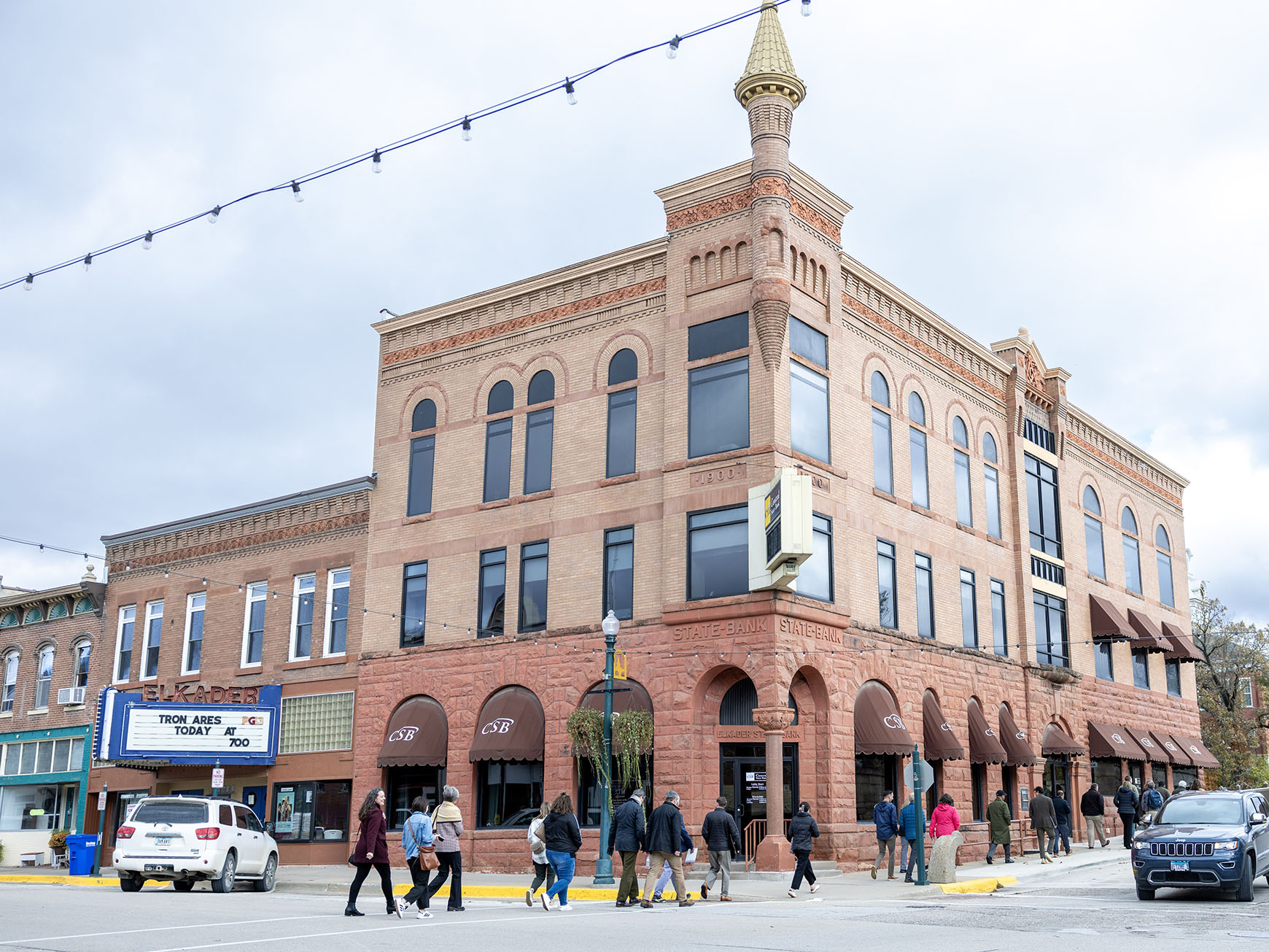 A group of people cross the street and walk up a sidewalk in front of a corner building.