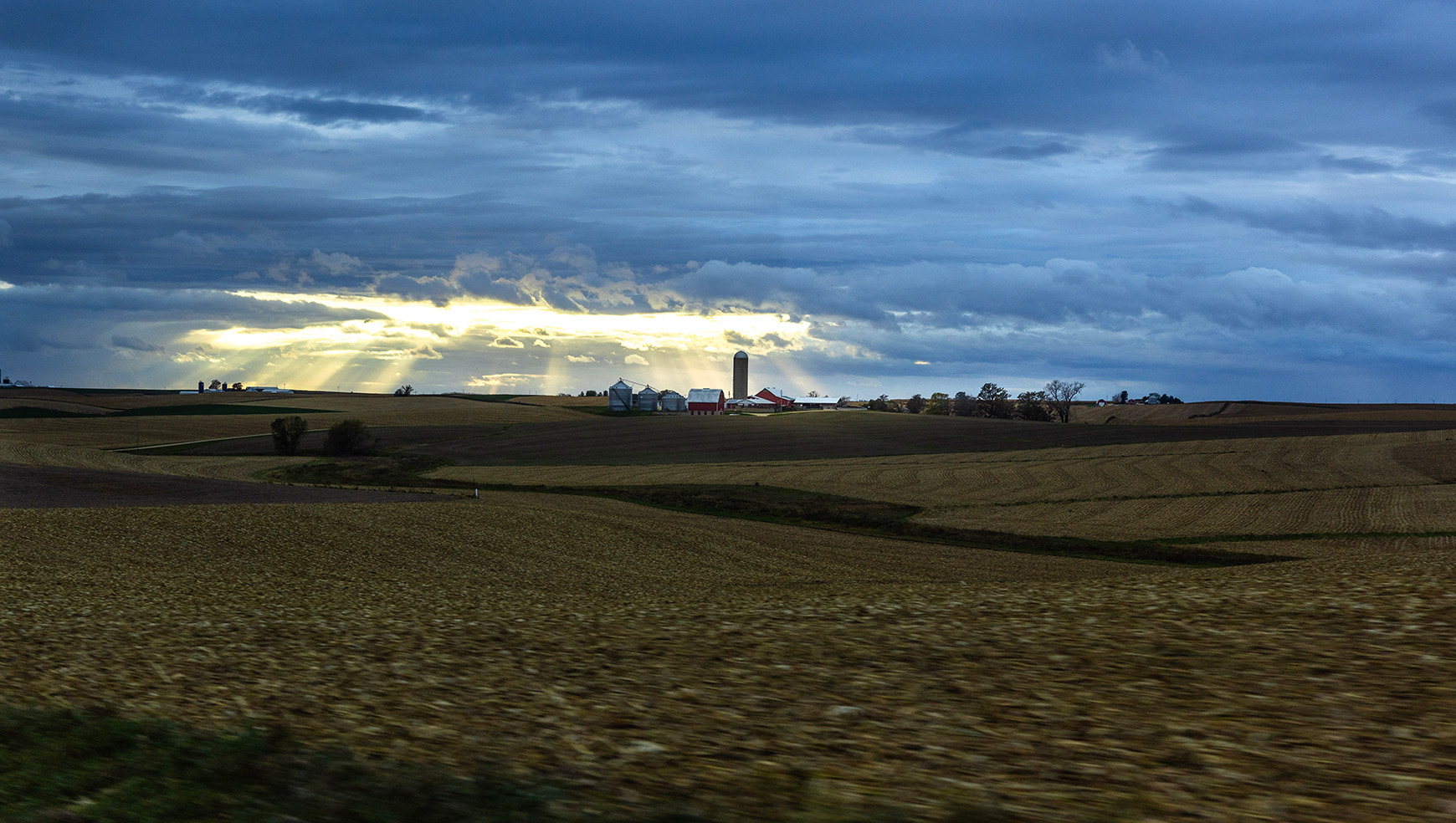 A cloudy sky opens up with rays of sunshine illuminating a barn, grain elevator, and rolling cornfield.